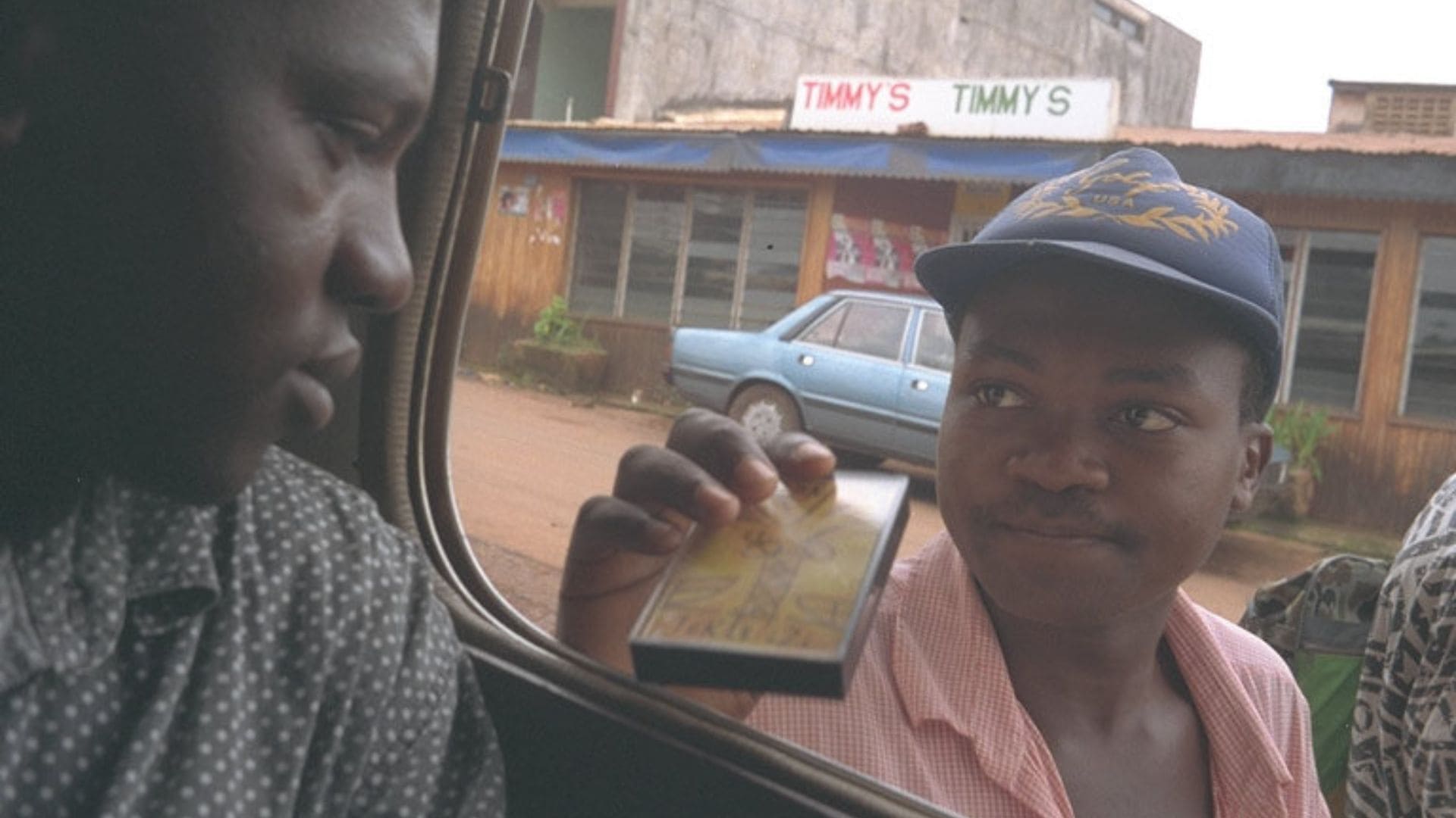 A man sitting in the back of a car looks out of the window towards a younger man showing him a small item.