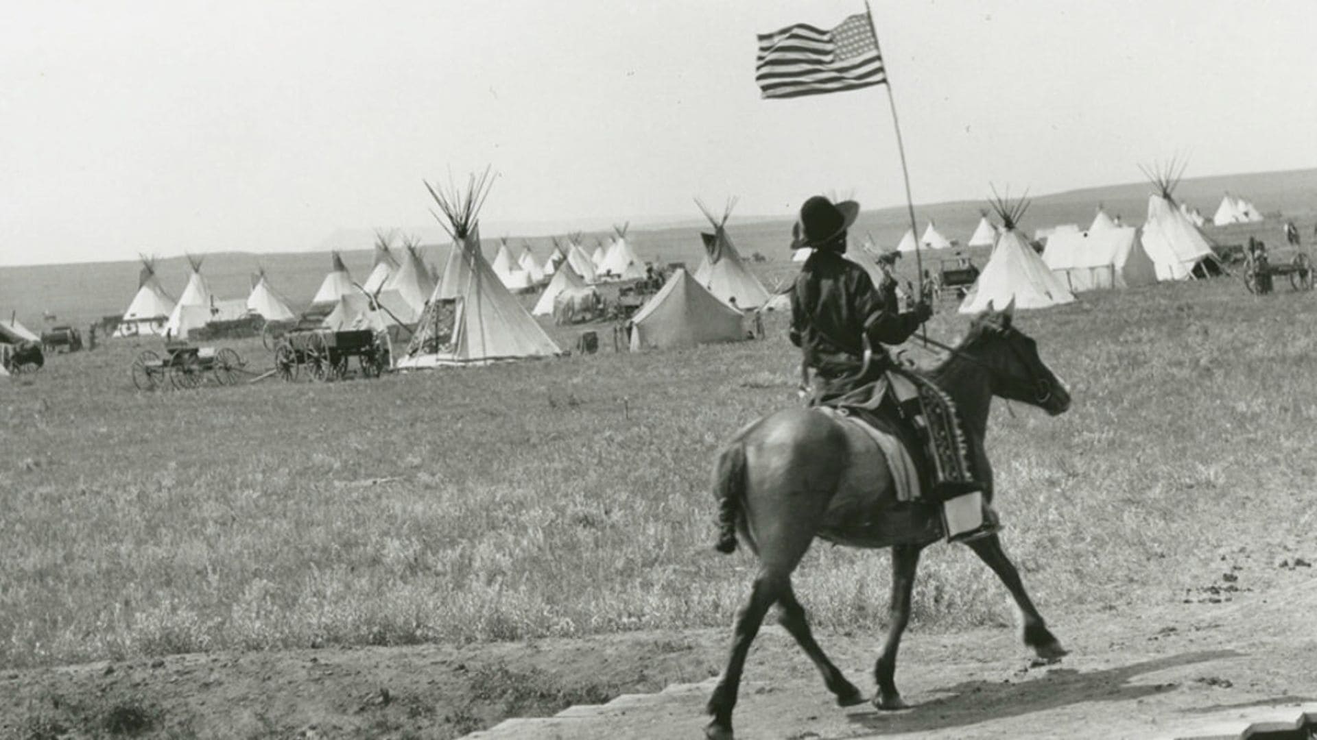 A man waves an American flag while riding his horse through a Native American village.
