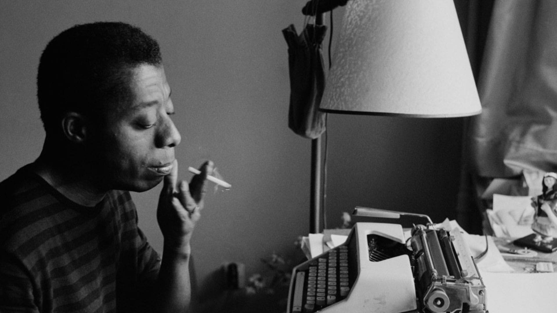 Author James Baldwin sits in front of a typewriter smoking a cigarette. 