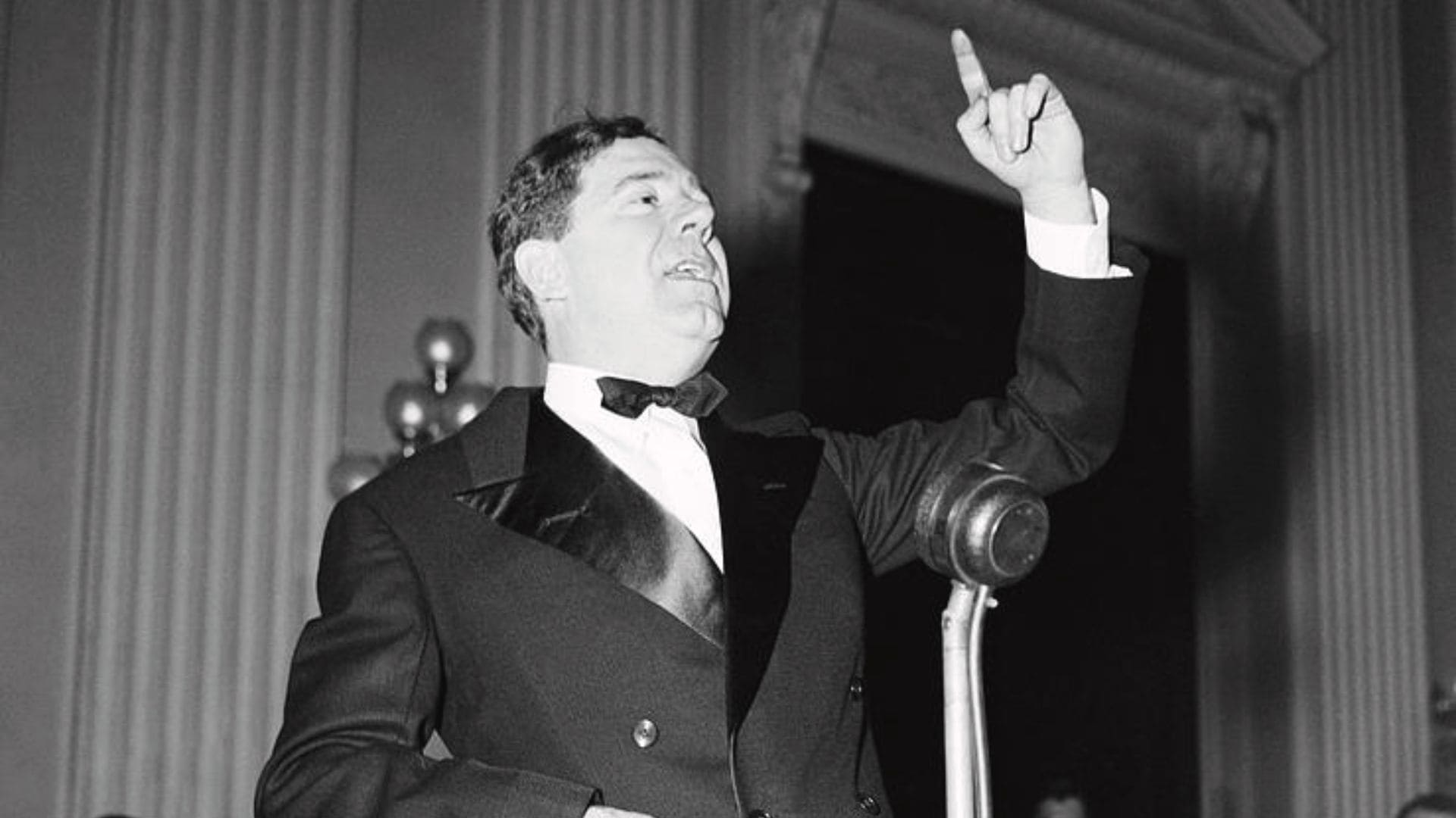 Politician Huey Long points his finger into the air while giving a speech in front of a podium.