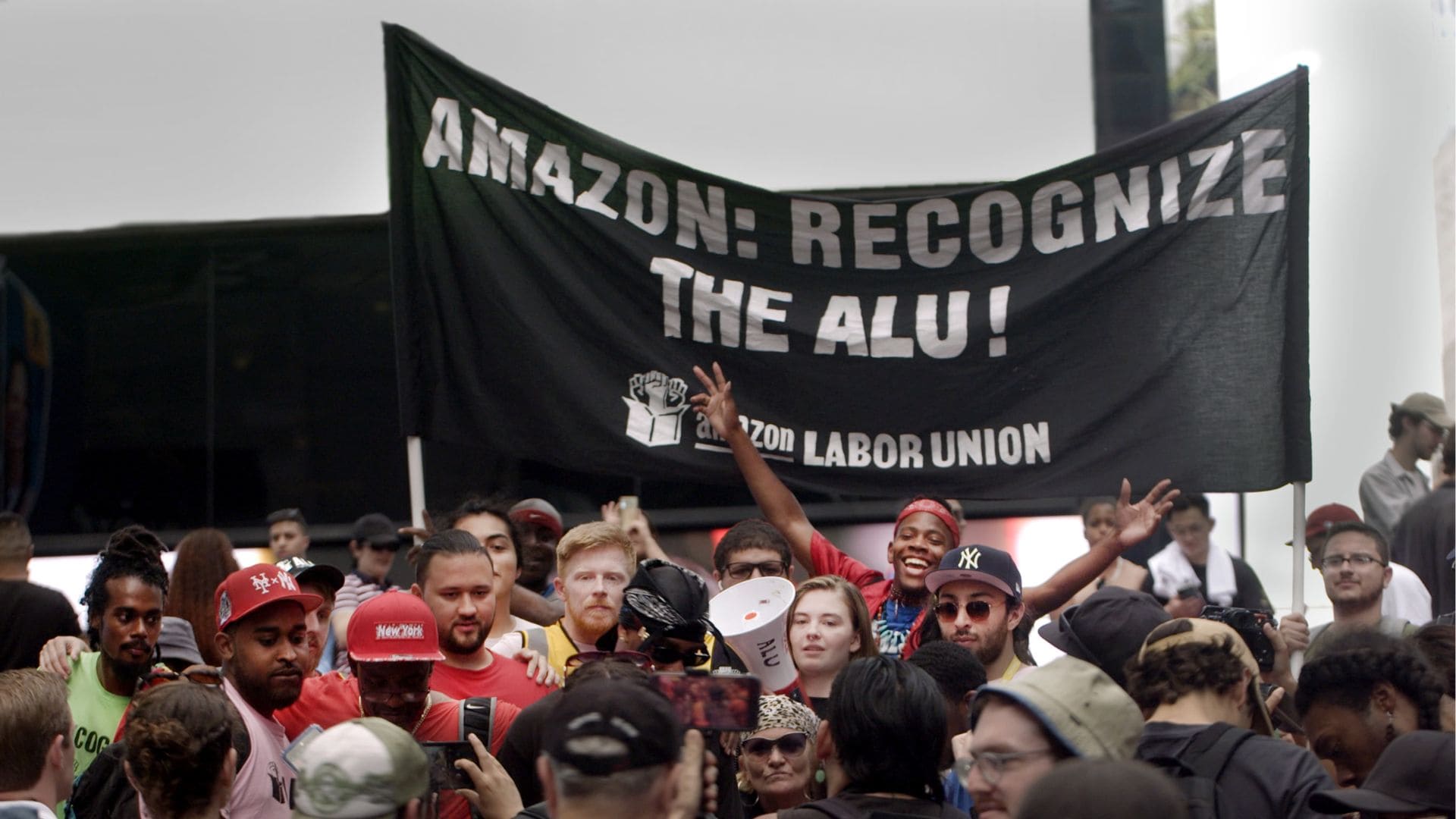 A crowd of people stand below a sign that reads: Amazon: Recognize the ALU!