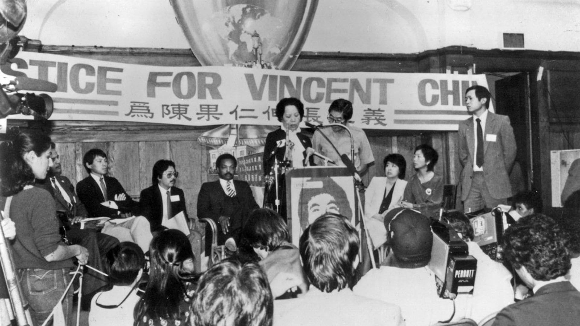 A woman gives a speech in front of a small, packed room.