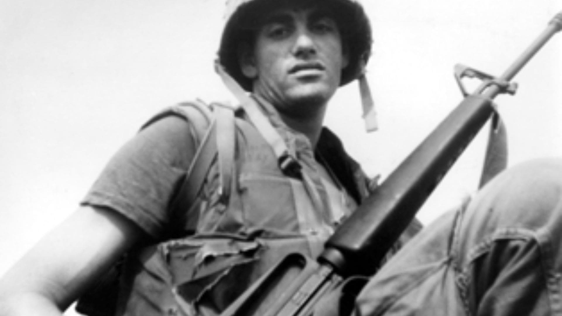 A man stands with a musket in protective military gear.