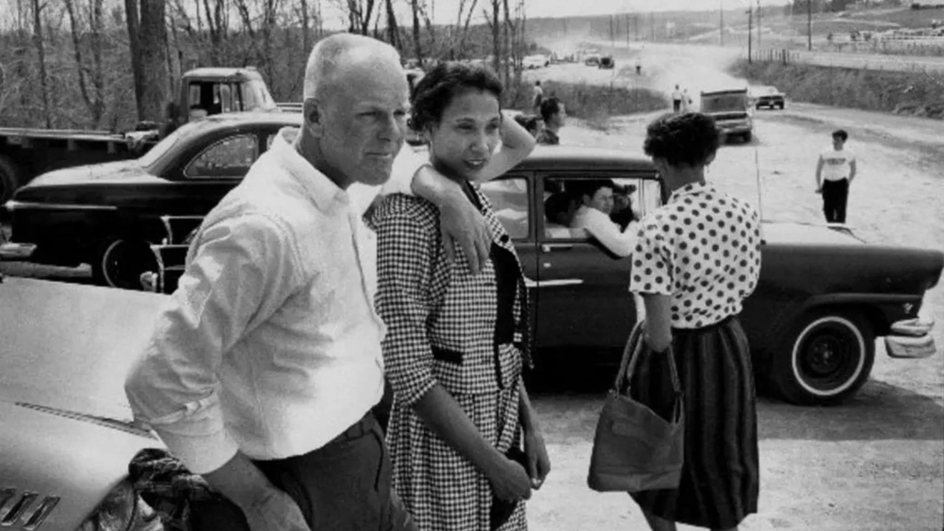 A couple stand together in front of their car in a parking lot.