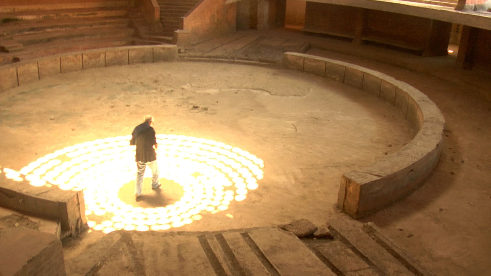 A man walks along the floor of an unfinished building, light shining through a skylight onto the ground.
