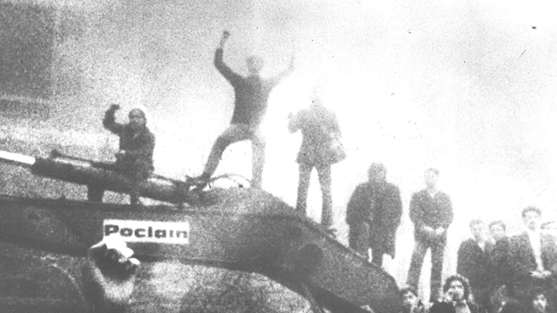 A small group of people stand on top of a machine, while fire or fog loom in the background.