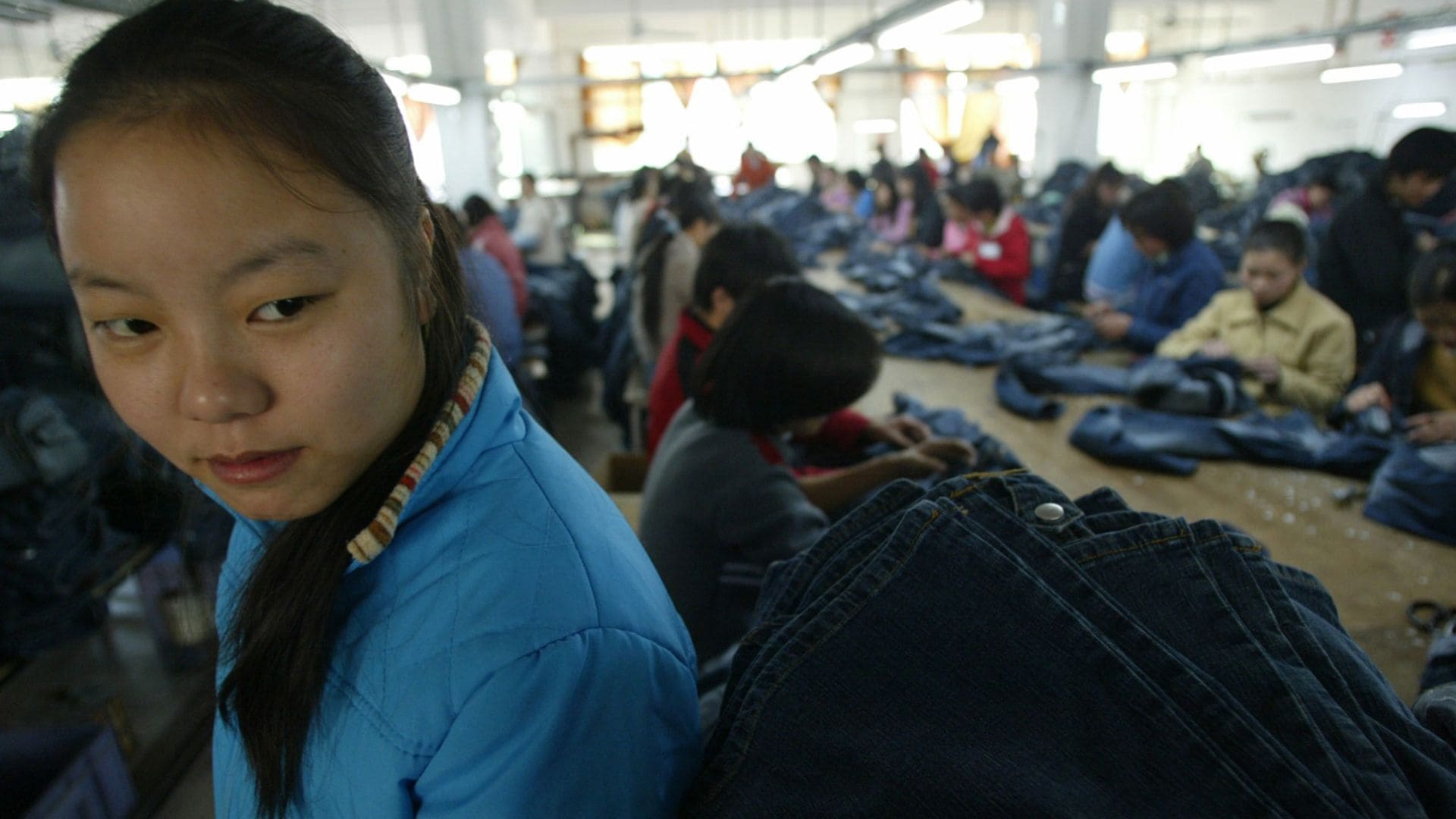 A girl smirks towards the camera, against a scene of children creating jeans at a factory.