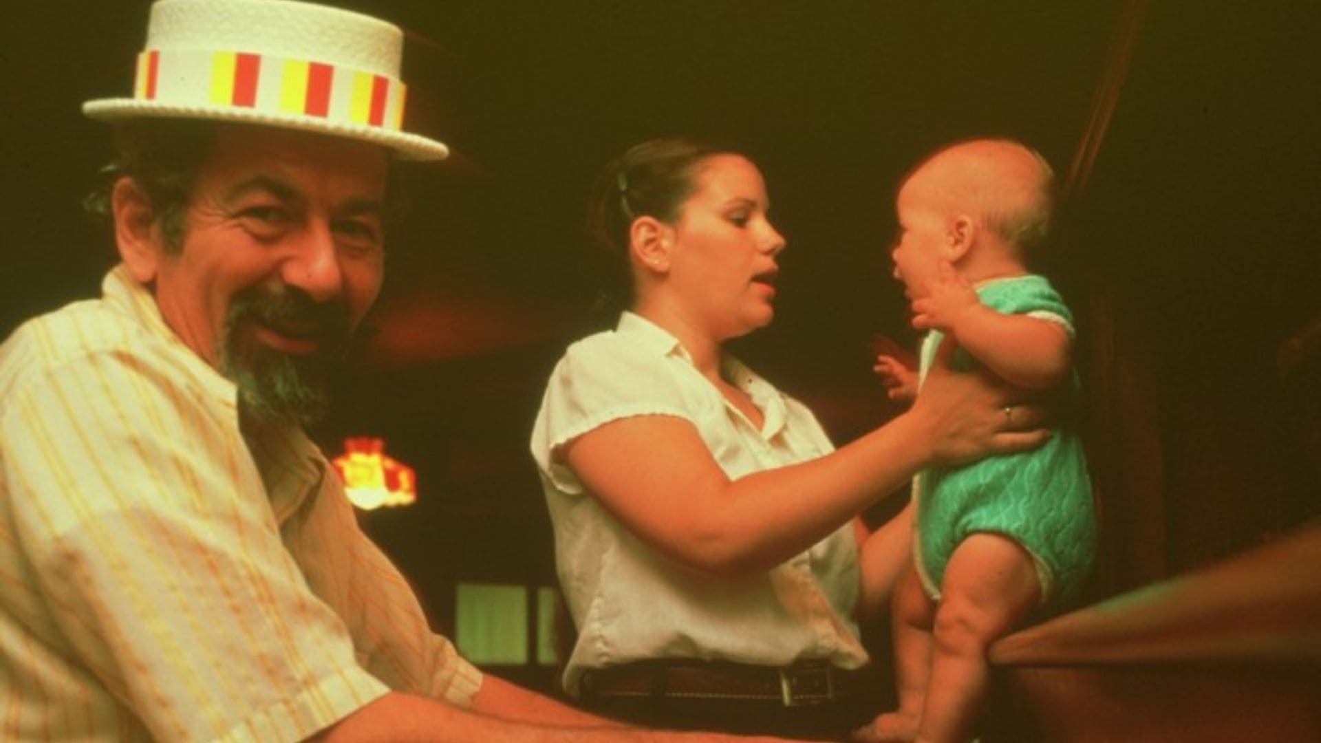 A man plays the piano while a woman stands behind him, holding their baby on the piano.