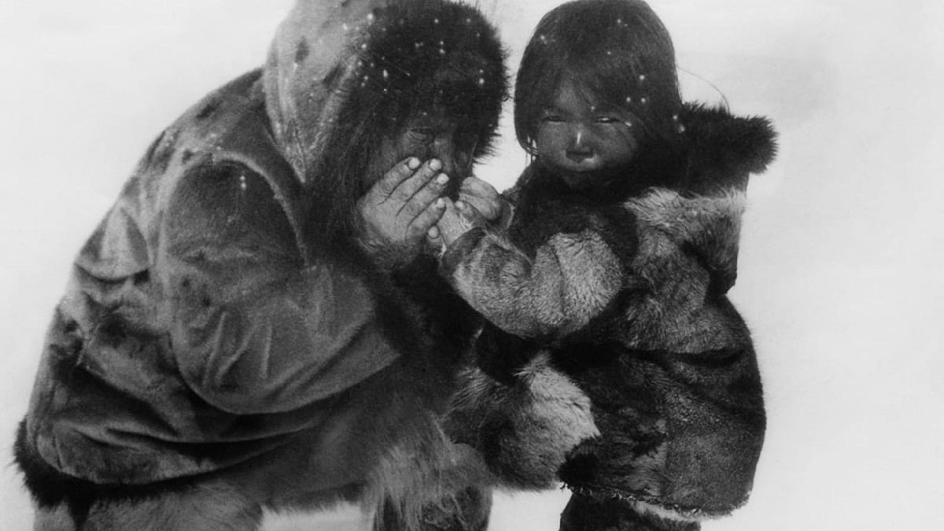 A woman and a child crouch in the snow, wearing heavy fur coats.