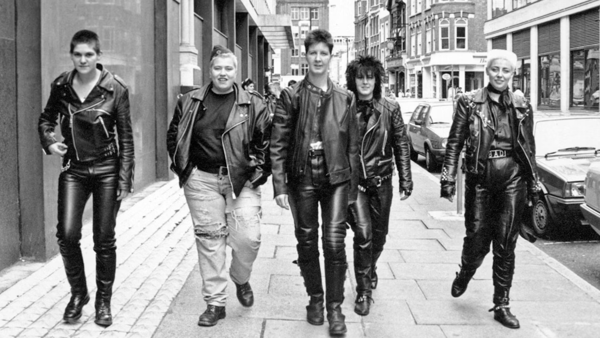 A group of women dressed in almost entirely leather outfits walk a city street.