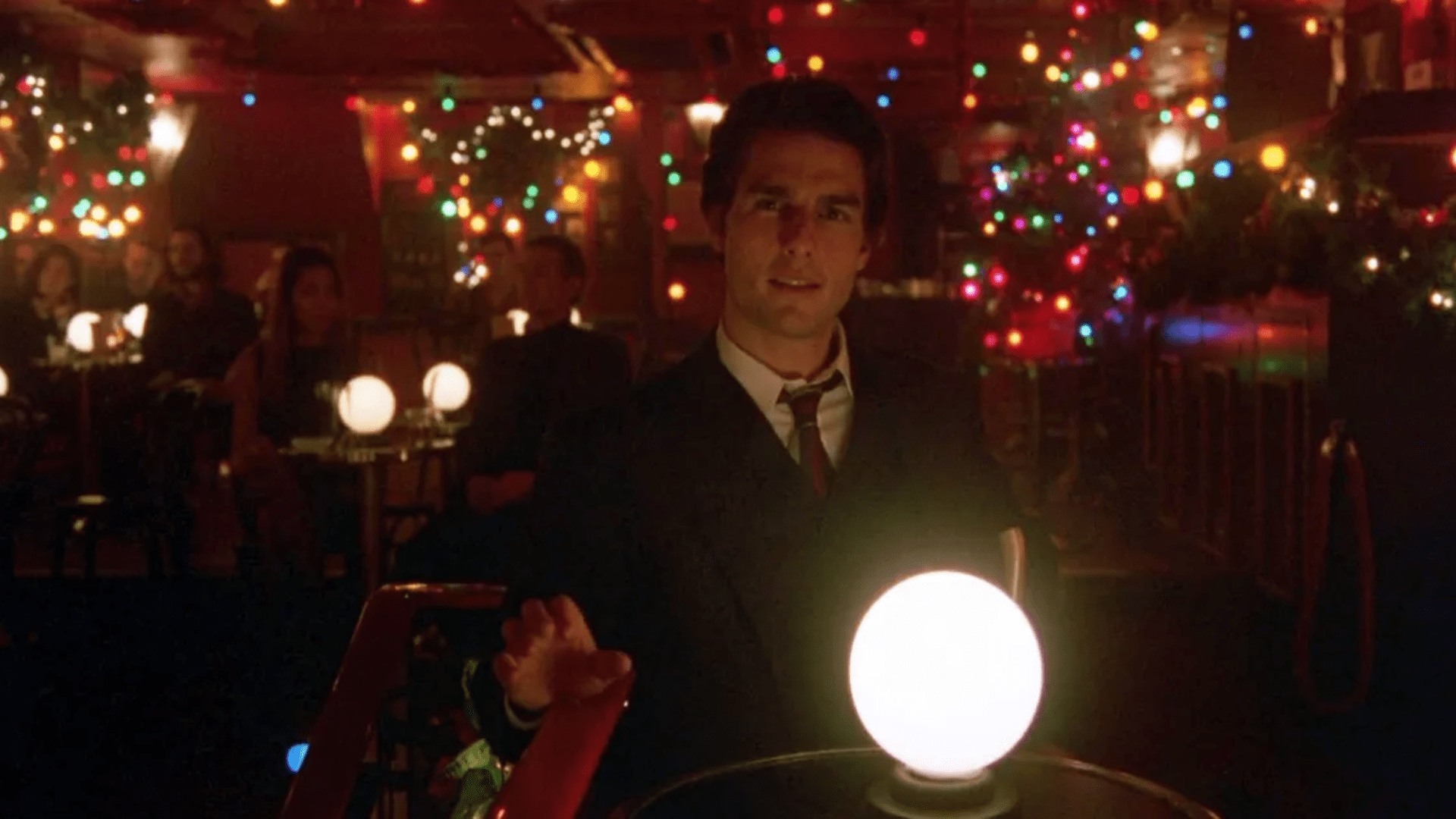 A man sits at a round table in a room dimly lit with holiday string lights.