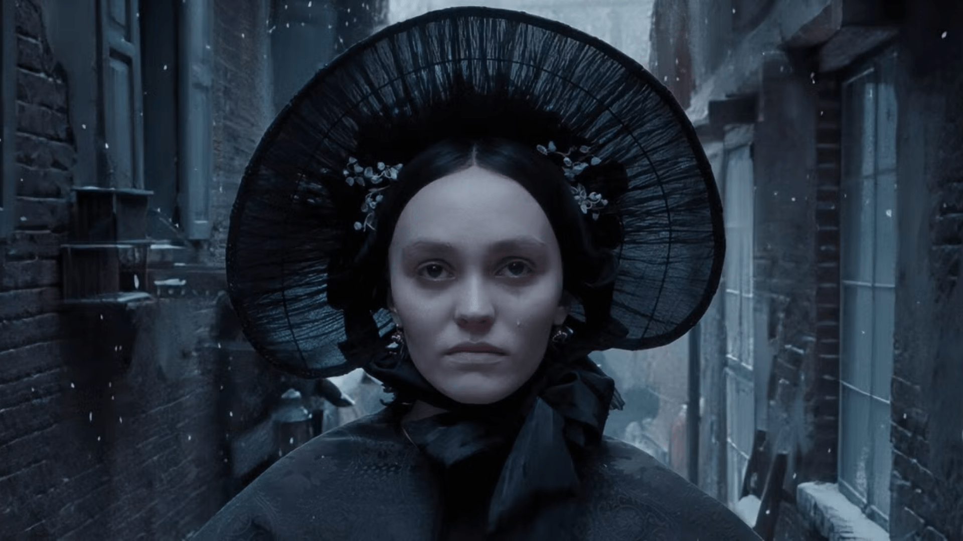 A woman dressed in black with a large, fanning hat looks straight at the viewer.