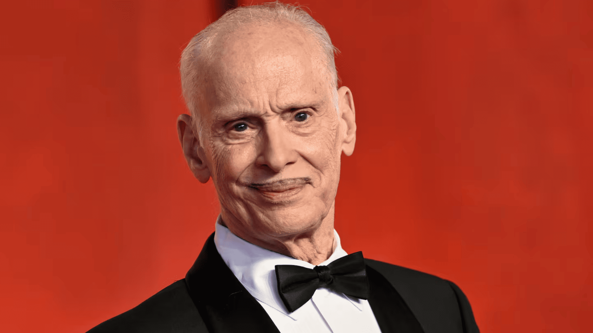A headshot of an older man in a suit, posing against a red backdrop.