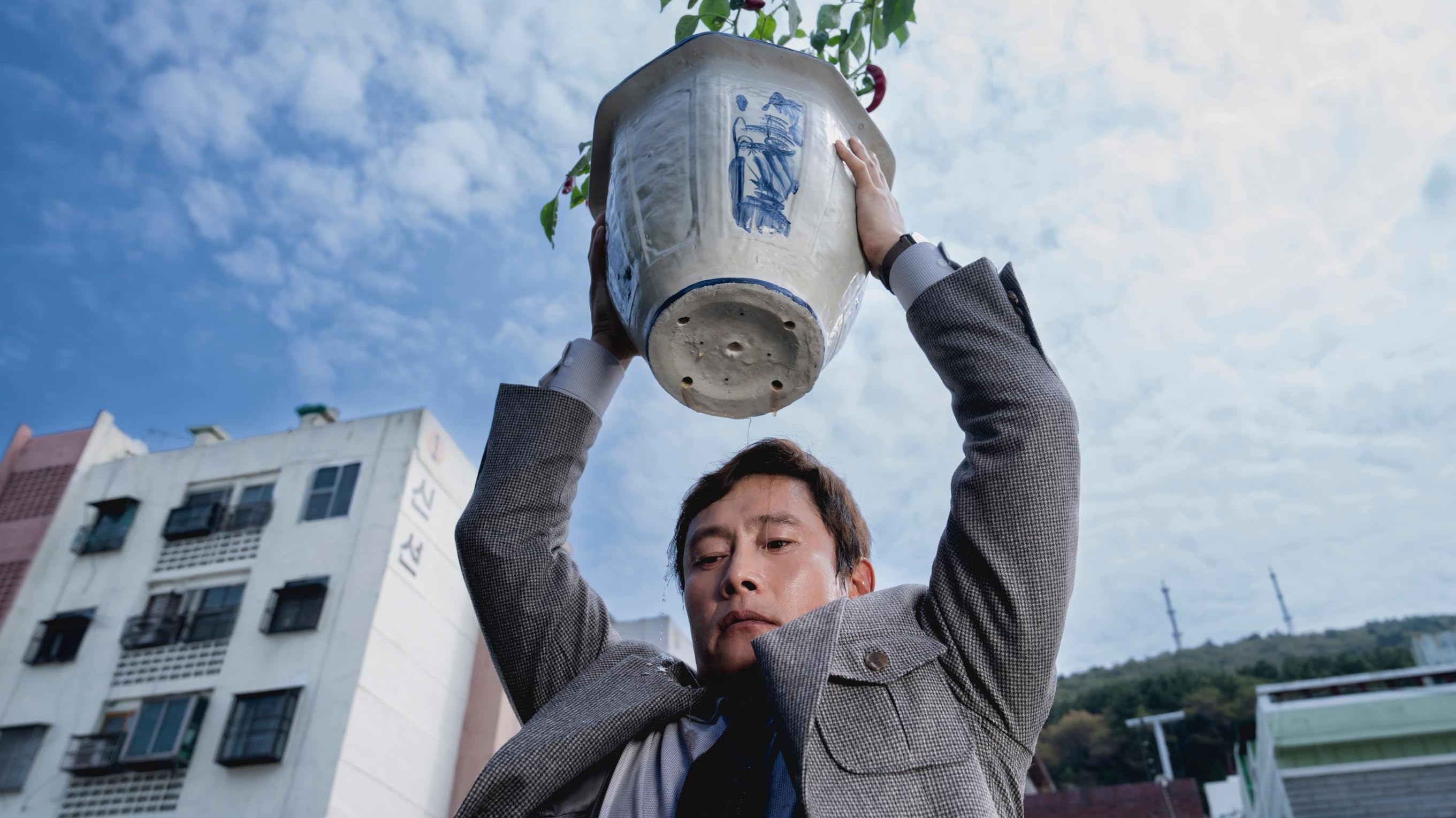 Lead actor Lee Byung-hun, as Man-su, holds a large potted plant above his head. Dirtied water leaks out of the pot's drainage holes onto his determined face. 