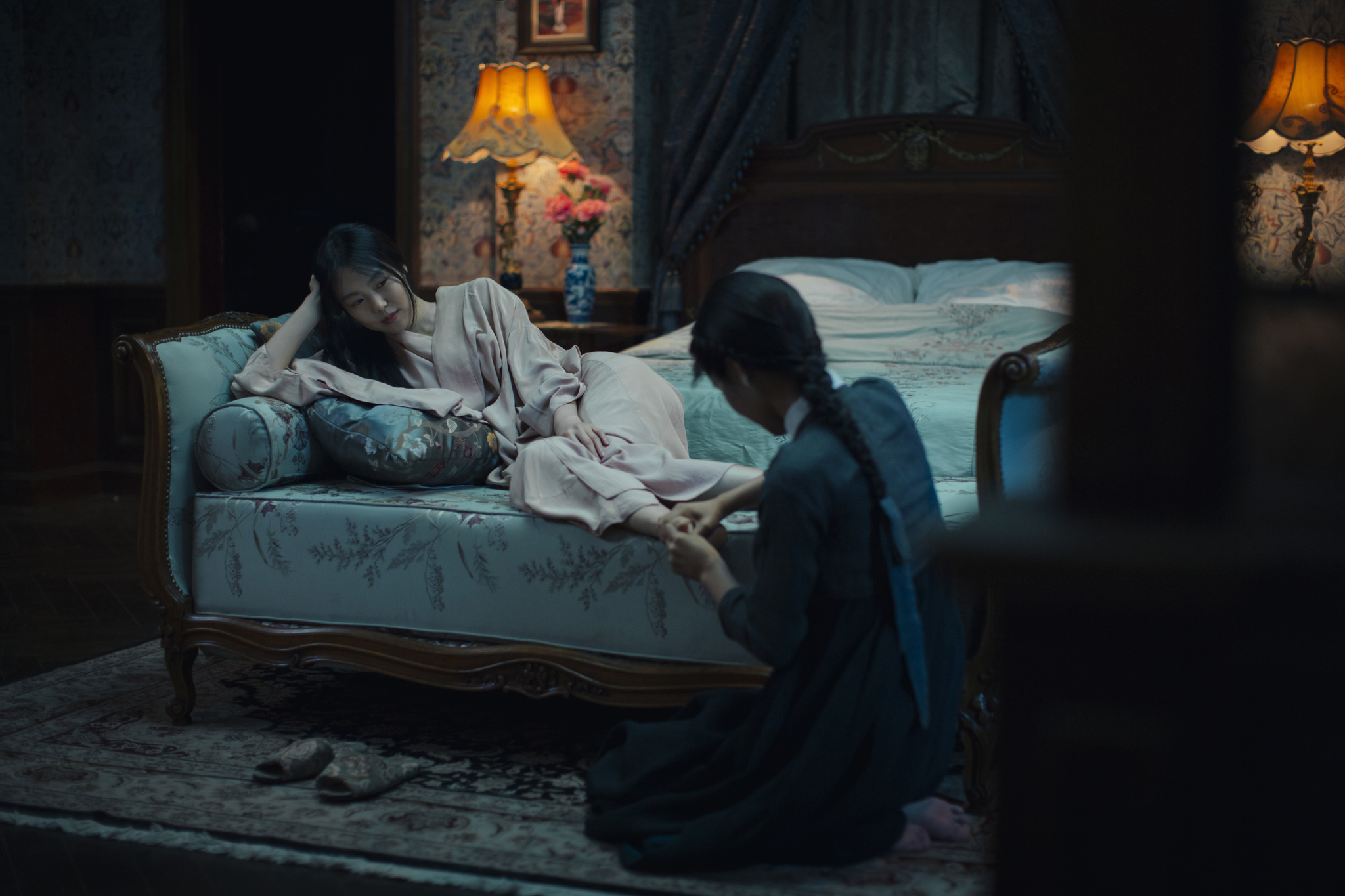 Lady Hideko (played by Kim Min-hee) comfortably lounges in a pink robe on an expensive-looking seat. She watches Sook-hee (Kim Tae-ri) kneeling on the ground in front of her as she massages Lady Hideko's feet. The room is dimly lit by table lamps behind them.