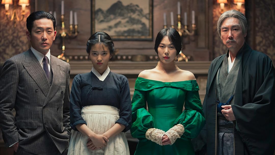 The main cast of The Handmaiden poses in front of the camera. They are dressed in 1930's period outfits in front of a fireplace decorated with golden candle holders and fancy wallpaper. Their expressions are intense and mysterious.