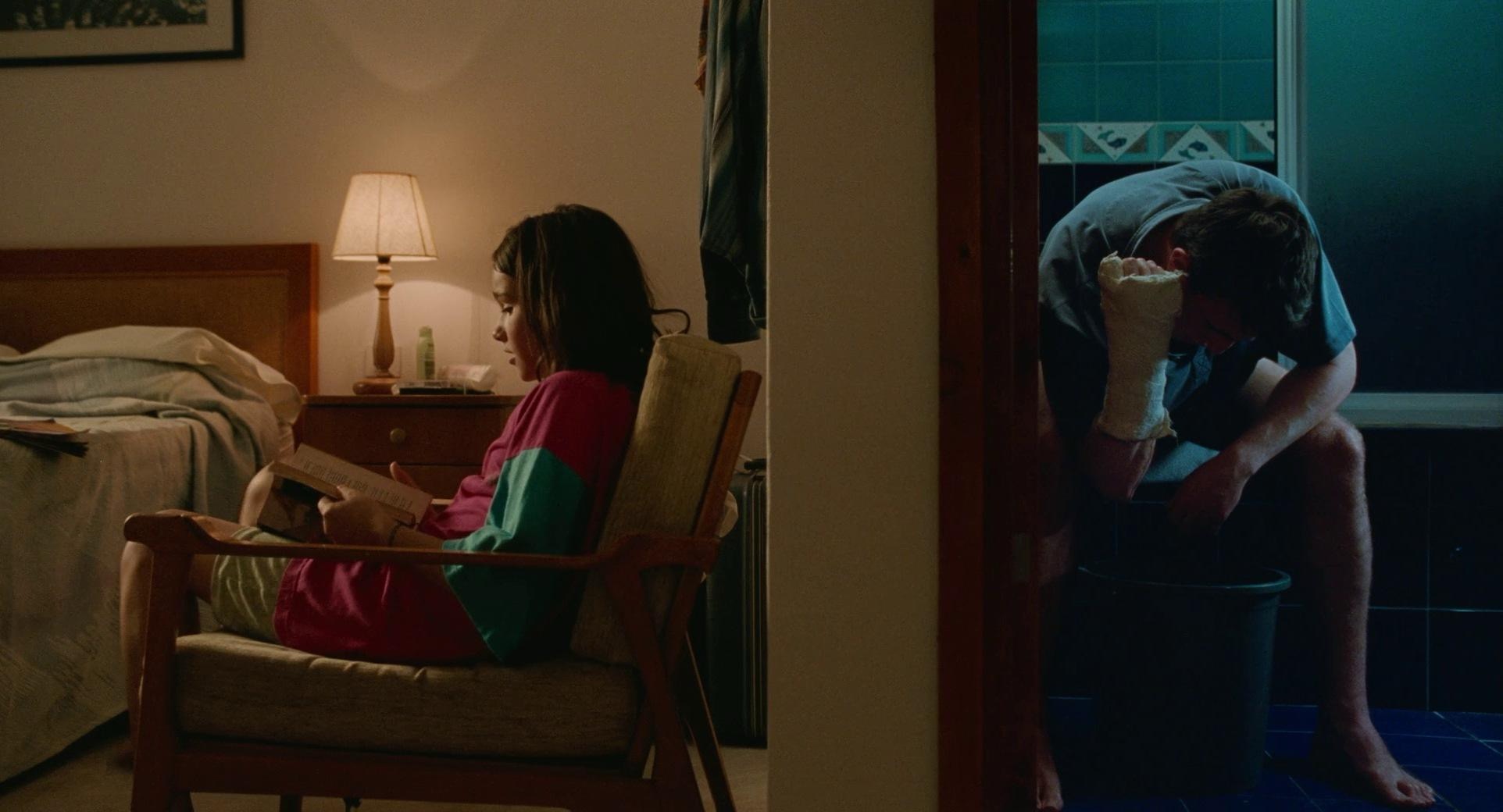 Scene from Aftersun where Sophie is sitting on one side of the wall in the hotel reading in a chair and Calum is on the other side in the bathroom looking down with a cast on his wrist