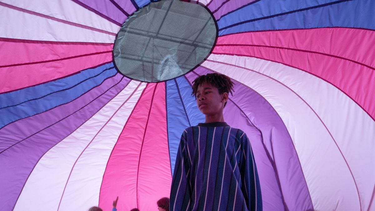 Scene from I Saw The TV Glow where young Owen is standing under a purple, pink & blue parachute